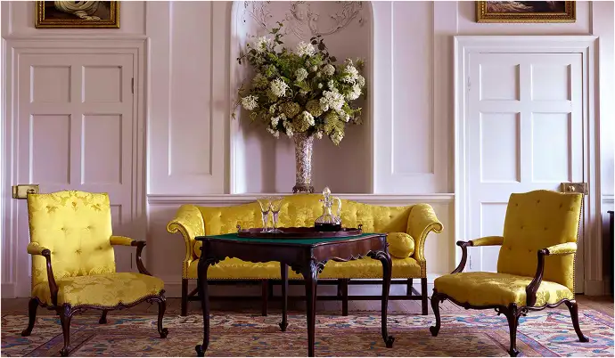A pale panelled drawing room furnished with a yellow silk sofa flanked by two matching armchairs, arranged around an antique games table set with glassware. A large arrangement of white hydrangeas sits in a silver vase in the alcove behind.