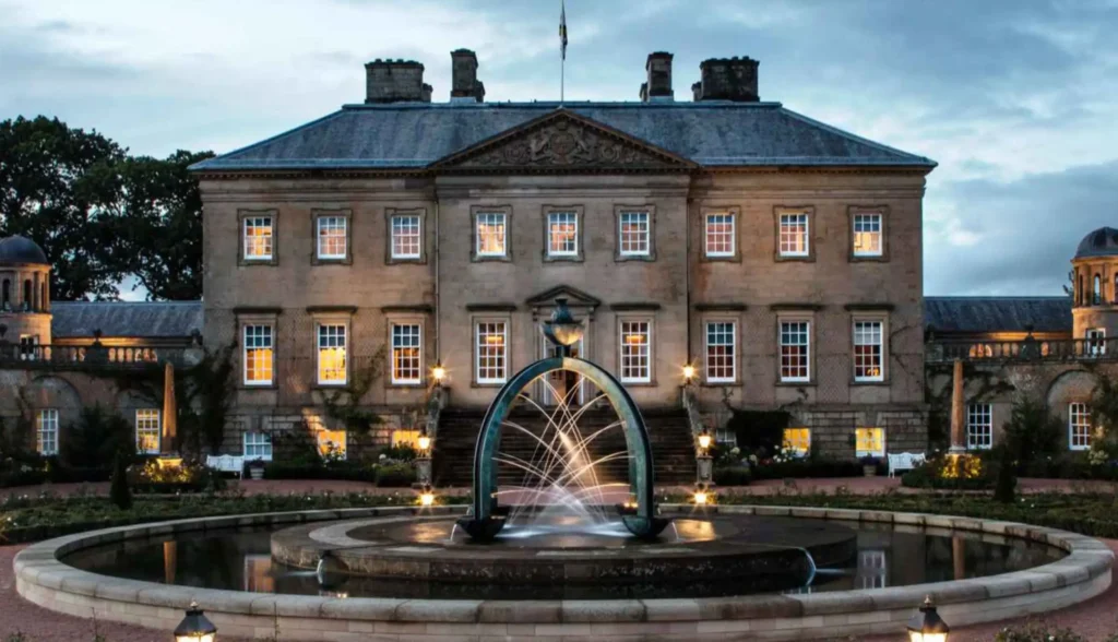 The Palladian front of Dumfries House at dusk, its sandstone facade lit from below and every window glowing warm gold against a blue-grey evening sky. In the foreground, the bronze arch fountain plays in its circular pool, with the formal rose garden and lantern-lit paths leading up to the entrance steps.