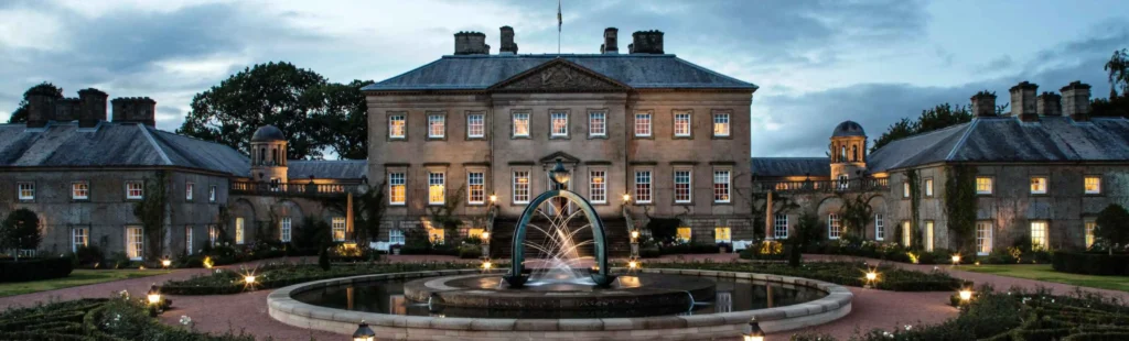 The Palladian front of Dumfries House at dusk, its sandstone facade lit from below and every window glowing warm gold against a blue-grey evening sky. In the foreground, the bronze arch fountain plays in its circular pool, with the formal rose garden and lantern-lit paths leading up to the entrance steps.