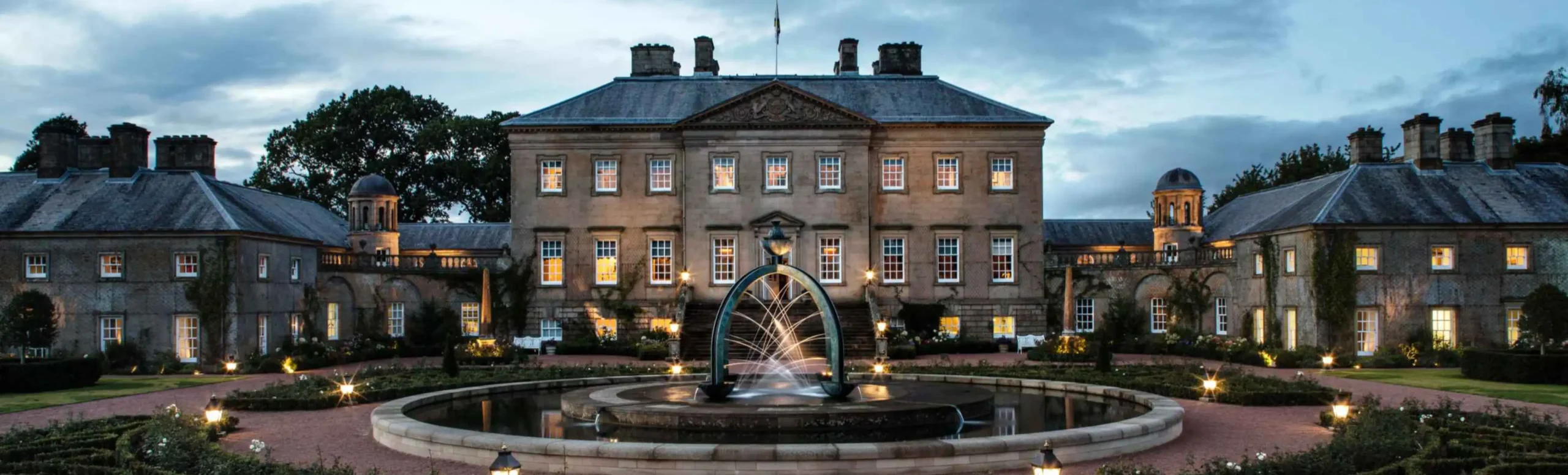 The Palladian front of Dumfries House at dusk, its sandstone facade lit from below and every window glowing warm gold against a blue-grey evening sky. In the foreground, the bronze arch fountain plays in its circular pool, with the formal rose garden and lantern-lit paths leading up to the entrance steps.