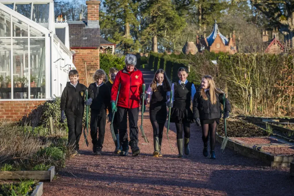 A garden volunteer in a red fleece leads a group of primary school pupils in uniform along a gravel path in a walled kitchen garden, each child carrying a garden fork. A Victorian glasshouse and raised beds line the path on a bright winter day.