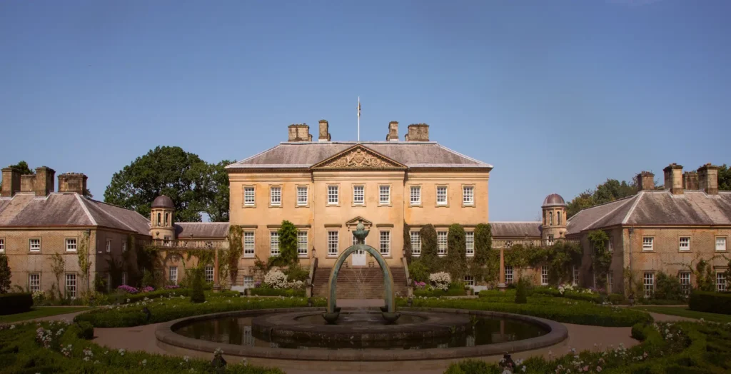 The symmetrical Palladian front of a honey-coloured sandstone country house with a carved pediment and flanking pavilions, seen across formal box-edged rose gardens and a circular pool with a bronze arch fountain at its centre.