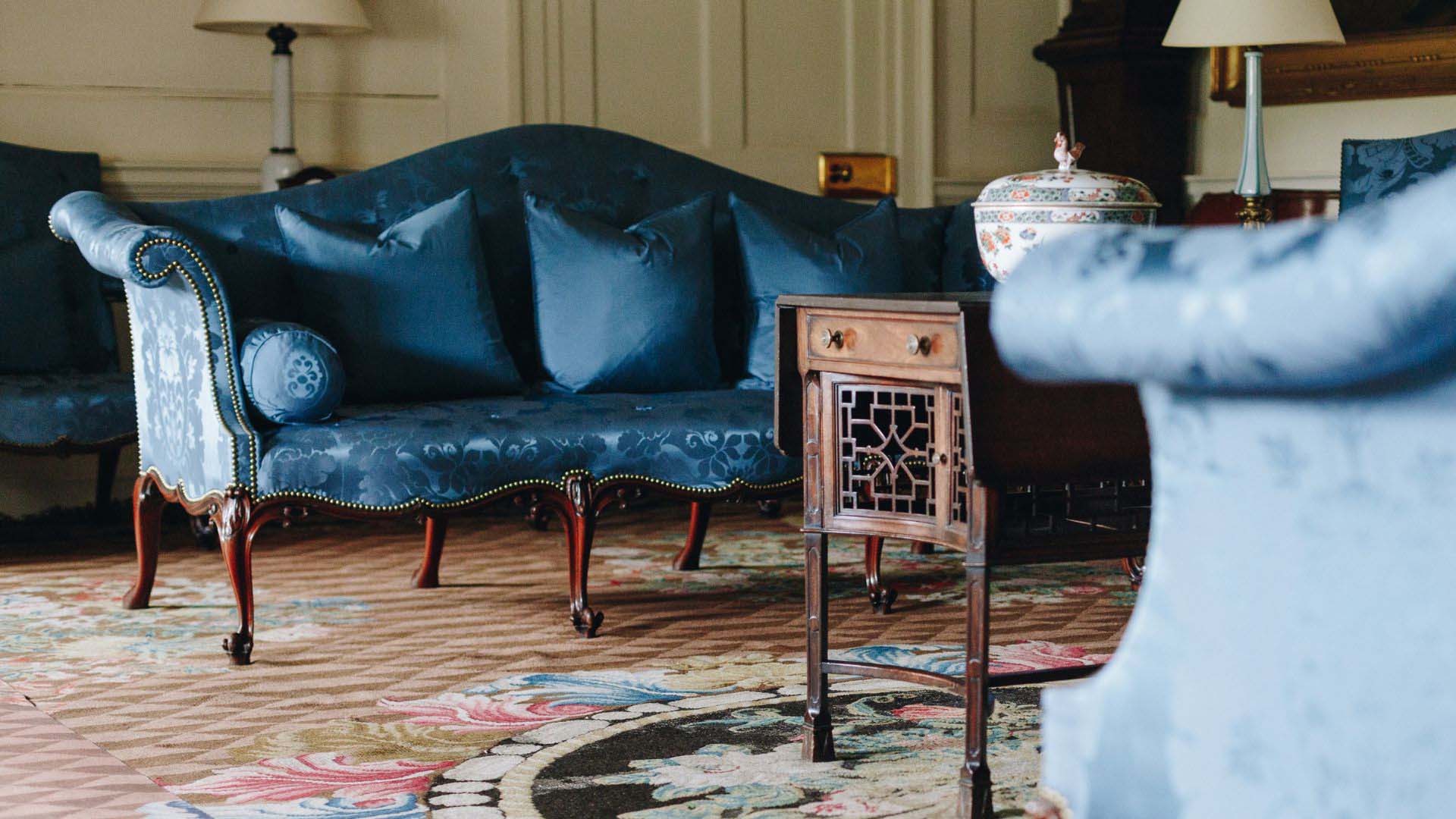 Low-angle view across a historic drawing room: a blue silk damask sofa with a serpentine back and carved mahogany cabriole legs, set with matching cushions and a bolster.
