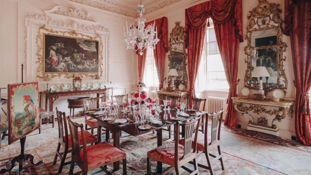 A grand dining room with ornate white plasterwork, deep red damask curtains and a Venetian glass chandelier