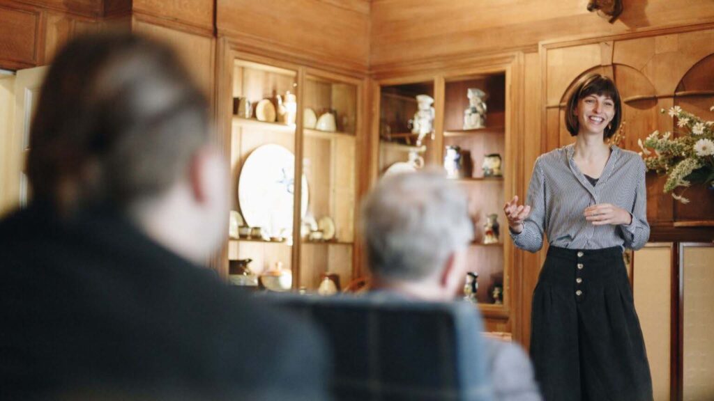 A guide in a striped shirt and dark culottes stands smiling and gesturing as she talks to visitors in a wood-panelled room lined with glazed display cabinets of ceramics and jugs. Two visitors are seen from behind in soft focus in the foreground, one seated.