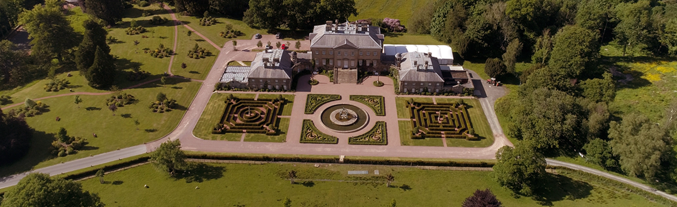 Aerial view of Dumfries house and its flanking pavilions, looking down on three formal parterres laid out below the south front — two symmetrical knot gardens either side of a central circular pool and fountain. Gravel paths, lawns, parkland and dense woodland surround the walled garden enclosure, with a marquee pitched beside the house.