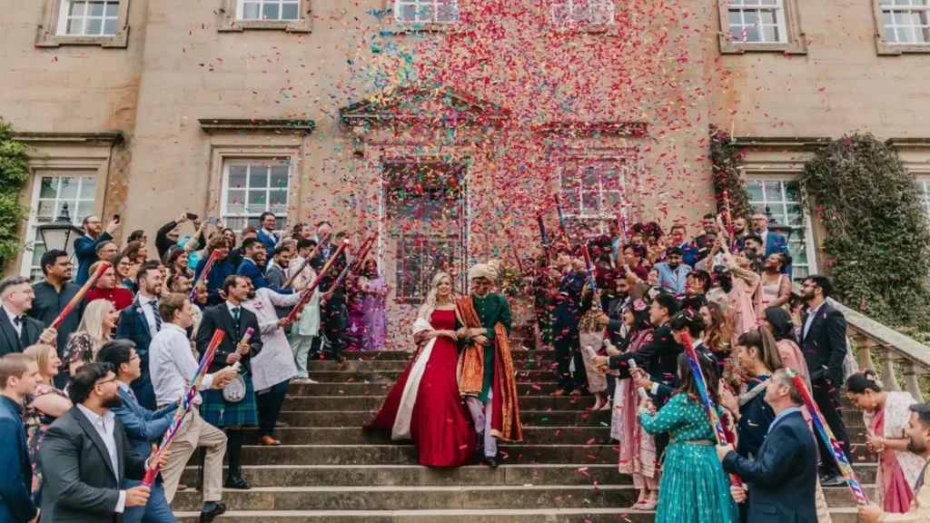 A newly married couple walk down the stone steps of a historic house as around a hundred guests lining both sides fire confetti cannons, filling the air with brightly coloured paper. The bride wears a red gown with a cream wrap; the groom wears a green and gold sherwani and turban.